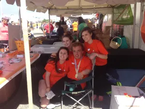 Team members in orange shirts laughing together under the booth tent, one wearing a medal