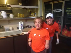 Two team members in orange shirts and gloves in a restaurant kitchen getting ready to bake