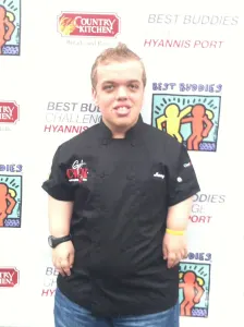 A young chef in a black chef's jacket in front of the Best Buddies Challenge backdrop