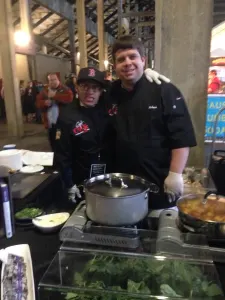 Two chefs in black jackets at a cooking station with a pot of food