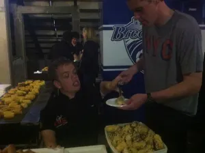 A chef in a black jacket serving food to a guest at the tailgate