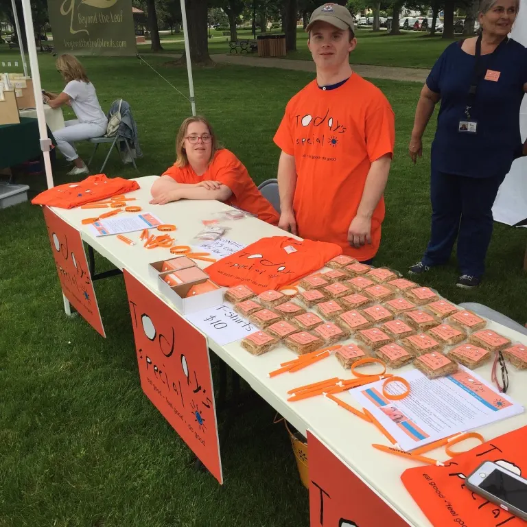 Two Today's Special team members in orange shirts at their booth table, covered in wrapped blondies, T-shirts, and Today's Special signs