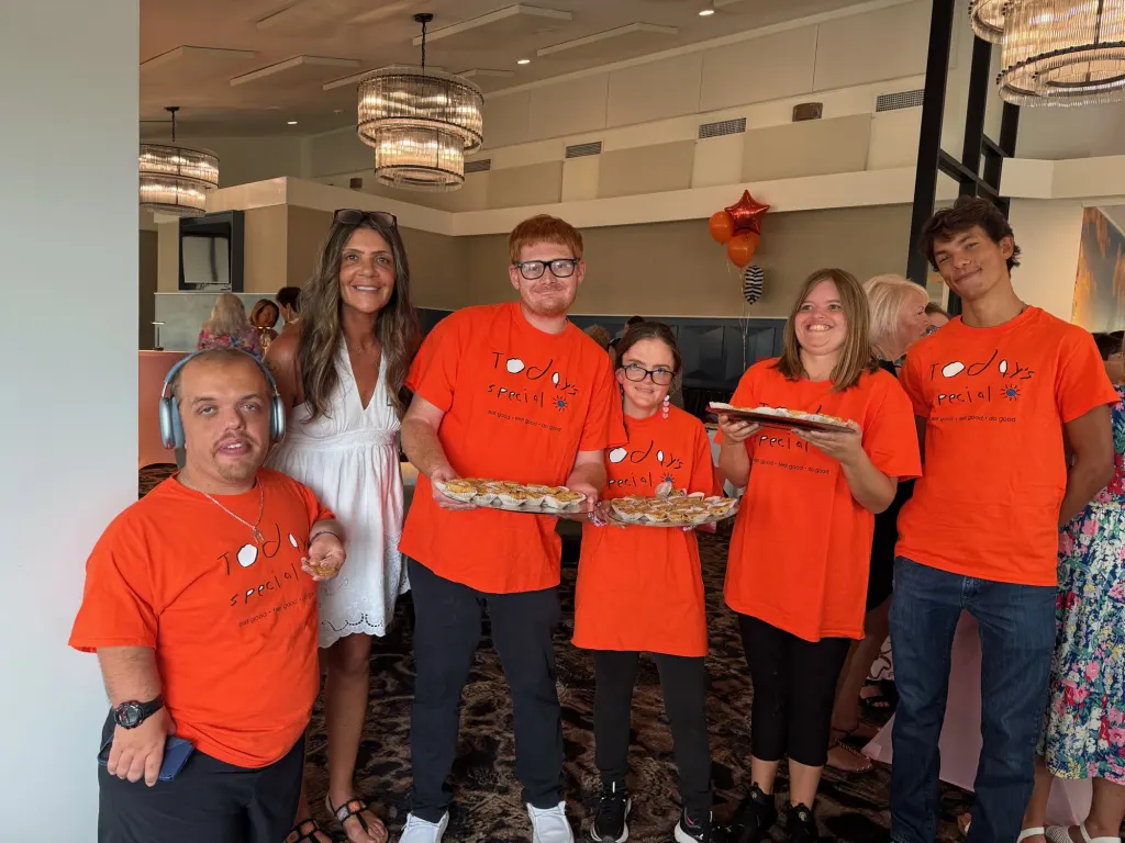 Four Today's Special team members in orange shirts holding trays of baked goods at an indoor event