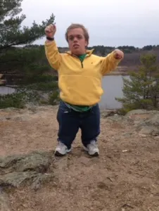 A young man in a yellow jacket cheering on a rocky overlook by a lake
