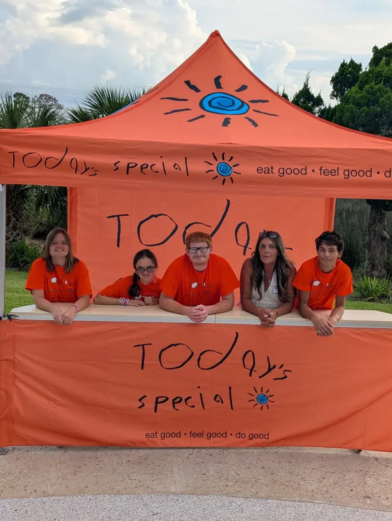 Five members of the Today's Special team in orange shirts lean on the counter of the orange Today's Special tent, under Harry's hand-drawn logo and sun