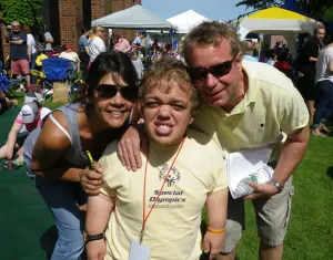 A smiling athlete in a Special Olympics shirt and medal, hugged by two supporters