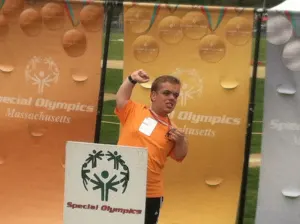 An athlete in an orange shirt raising a fist in front of Special Olympics Massachusetts banners