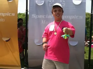 A smiling athlete in a pink shirt showing a medal in front of a Special Olympics banner